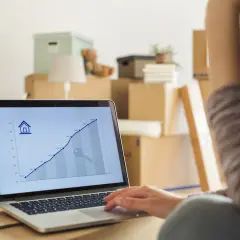 Na ekranie laptopa wykres przedstawiający wzrost cen nieruchomości, przypominający stopę referencyjną NBP. W tle kartony do przeprowadzki.