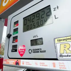 Cyfrowy wyświetlacz dystrybutora paliwa pokazuje cenę 4,29 zł za litr. Obok widnieje informacja o promocji "Co 6. kawa gratis", a na dole znajduje się naklejka z sercem i napisem "Świątecznej pomocy". W tle widać fragment samochodu i drzewa. To nie jes...