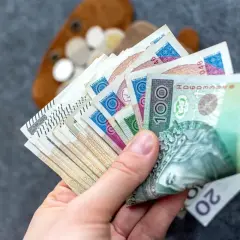 Dłoń trzyma polskie banknoty, w tym 100 zł. W tle widać portfel z monetami. Czy to wystarczy na najniższą rentę netto?