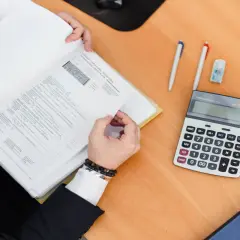 Osoba w garniturze przegląda dokumenty, obok kalkulator i długopisy. Czy to rozliczenie podatkowe, czy może ebit co to?
