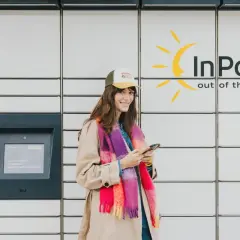 Uśmiechnięta kobieta z telefonem stoi przy paczkomacie InPost. Wygodne akcje InPost to jej codzienność.