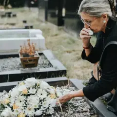 Kobieta w żałobie klęczy przy grobie, ocierając łzy. Obok leży bukiet kwiatów. Zastanawia się, od kiedy wzrośnie zasiłek pogrzebowy.