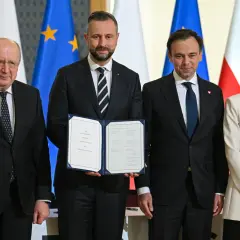 Cztery osoby stoją przed flagami. Jedna z nich trzyma dokument "SAFE LOAN AGREEMENT".
