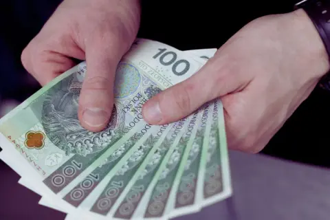 Dłonie trzymają stos banknotów 100 zł. To może być wypłata, premia lub kwota bliska minimalna brutto.