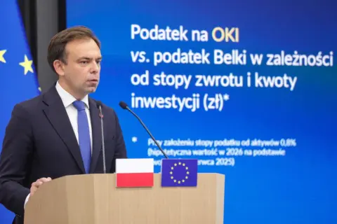 Mężczyzna w garniturze przemawia na tle ekranu z napisem "Podatek na OKI vs. podatek Belki". Dyskusja o zmianach w podatkach.