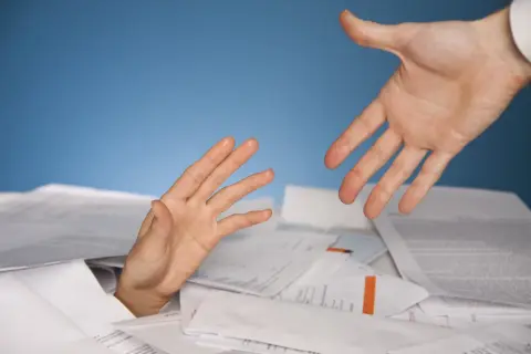 Dłoń wyciągnięta z papierów, druga dłoń oferuje pomoc. Symbolizuje trudności i potrzebę wsparcia w procesie restrukturyzacji.