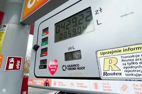 Cyfrowy wyświetlacz dystrybutora paliwa pokazuje cenę 4,29 zł za litr. Obok widnieje informacja o promocji "Co 6. kawa gratis", a na dole znajduje się naklejka z sercem i napisem "Świątecznej pomocy". W tle widać fragment samochodu i drzewa. To nie jes...