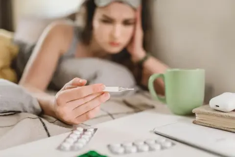 Chora dziewczyna z termometrem w ręku, leżąca w łóżku. Obok leki i kubek herbaty. L4 online 49 zł.