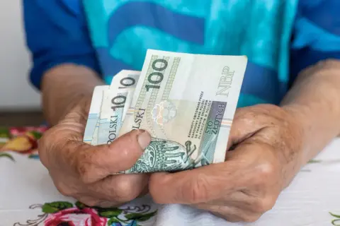 Dłonie starszej osoby trzymają banknoty 100 zł. Czy to wystarczy na najniższą emeryturę 2026?