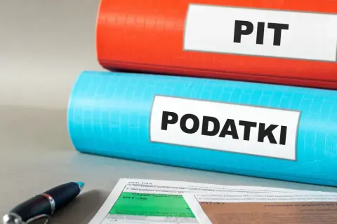 Dwa segregatory, jeden czerwony z napisem "PIT", drugi niebieski z napisem "PODATKI", leżą na biurku obok formularzy podatkowych i długopisu.