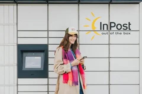Uśmiechnięta kobieta z telefonem stoi przy paczkomacie InPost. Wygodne akcje InPost to jej codzienność.