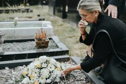 Kobieta w żałobie klęczy przy grobie, ocierając łzy. Obok leży bukiet kwiatów. Zastanawia się, od kiedy wzrośnie zasiłek pogrzebowy.