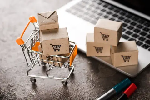 Miniaturowy wózek sklepowy z paczkami na tle laptopa symbolizuje e-commerce i wygodne zakupy online.