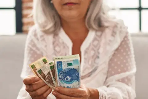 Kobieta z siwymi włosami trzyma polskie banknoty. To symbol oszczędności na emeryturę kapitałową dla kobiet.