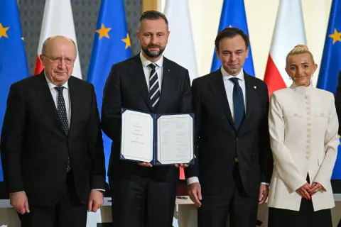 Cztery osoby stoją przed flagami. Jedna z nich trzyma dokument "SAFE LOAN AGREEMENT".