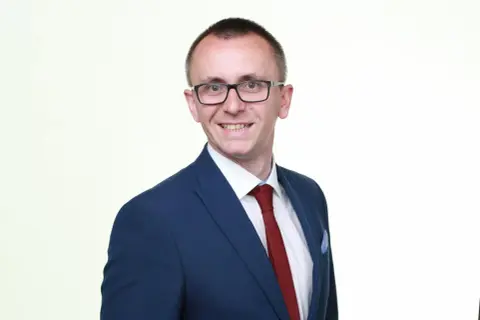 Mężczyzna w okularach, garniturze i krawacie, uśmiecha się na białym tle.