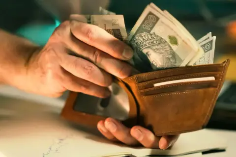 Dłonie wkładają polskie banknoty do brązowego portfela. Może to być pomoc w opłaceniu rachunków, np. bon ciepłowniczy.
