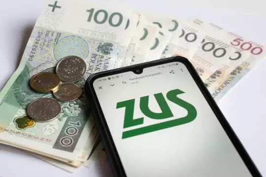 Złotówki i monety obok telefonu z logo ZUS. Może to symbolizować starania o zasiłek opiekuńczy.
