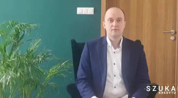 Mężczyzna w garniturze siedzi w fotelu obok rośliny, na tle zielonej ściany i drewnianych drzwi.