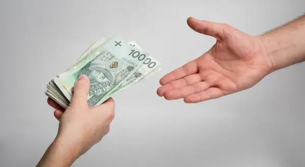 Dłoń podaje plik banknotów 100 zł. Zastanawiasz się, ile wynoszą odsetki od niezapłaconej faktury?
