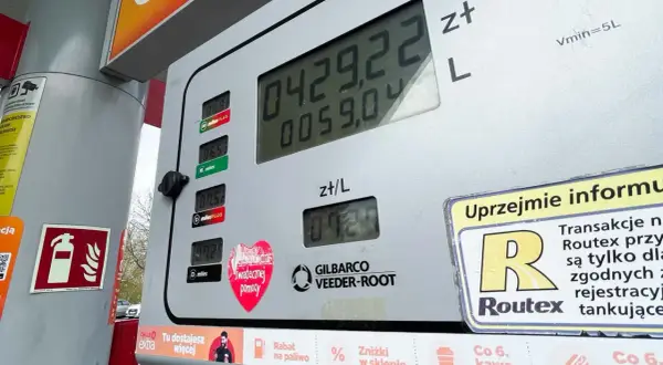 Cyfrowy wyświetlacz dystrybutora paliwa pokazuje cenę 4,29 zł za litr. Obok widnieje informacja o promocji "Co 6. kawa gratis", a na dole znajduje się naklejka z sercem i napisem "Świątecznej pomocy". W tle widać fragment samochodu i drzewa. To nie jes...