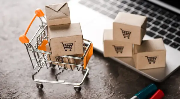 Miniaturowy wózek sklepowy z paczkami na tle laptopa symbolizuje e-commerce i wygodne zakupy online.