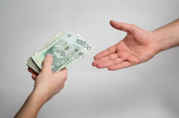 Dłoń podaje plik banknotów 100 zł. Zastanawiasz się, ile wynoszą odsetki od niezapłaconej faktury?