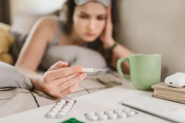 Chora dziewczyna z termometrem w ręku, leżąca w łóżku. Obok leki i kubek herbaty. L4 online 49 zł.