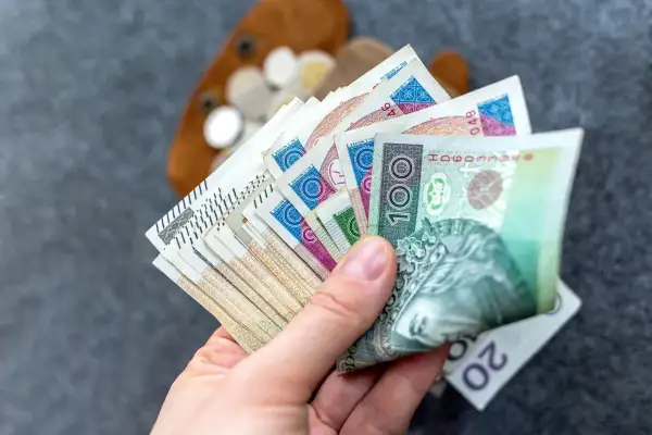 Dłoń trzyma polskie banknoty, w tym 100 zł. W tle widać portfel z monetami. Czy to wystarczy na najniższą rentę netto?