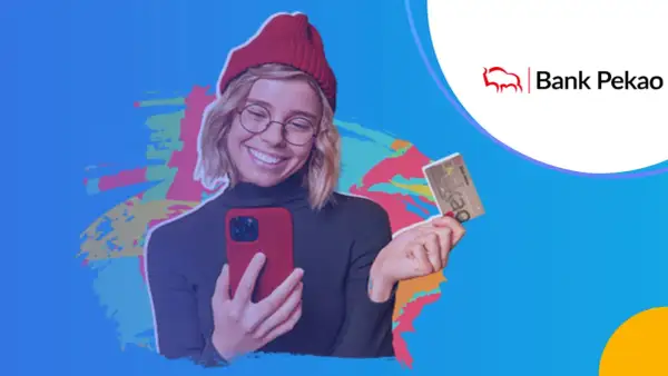 Uśmiechnięta młoda kobieta w czerwonej czapce trzyma telefon i kartę bankową Pekao.