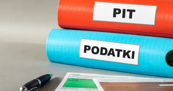 Dwa segregatory, jeden czerwony z napisem "PIT", drugi niebieski z napisem "PODATKI", leżą na biurku obok formularzy podatkowych i długopisu.