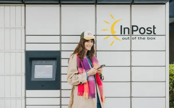Uśmiechnięta kobieta z telefonem stoi przy paczkomacie InPost. Wygodne akcje InPost to jej codzienność.