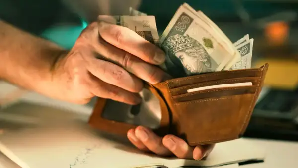 Dłonie wkładają polskie banknoty do brązowego portfela. Może to być pomoc w opłaceniu rachunków, np. bon ciepłowniczy.